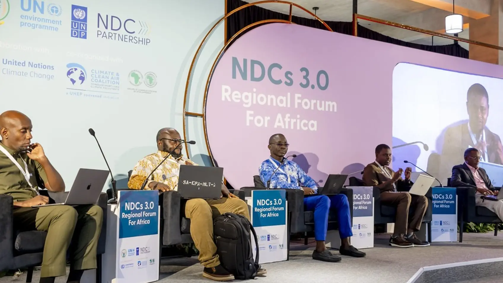 A panel discussion at the NDCs 3.0 Regional Forum for Africa. Four panelists are seated on stage, with the event's logo displayed behind them. The man in the center, wearing a patterned shirt, is speaking into a microphone, while the others listen attentively. The audience, visible in the foreground, is focused on the discussion.