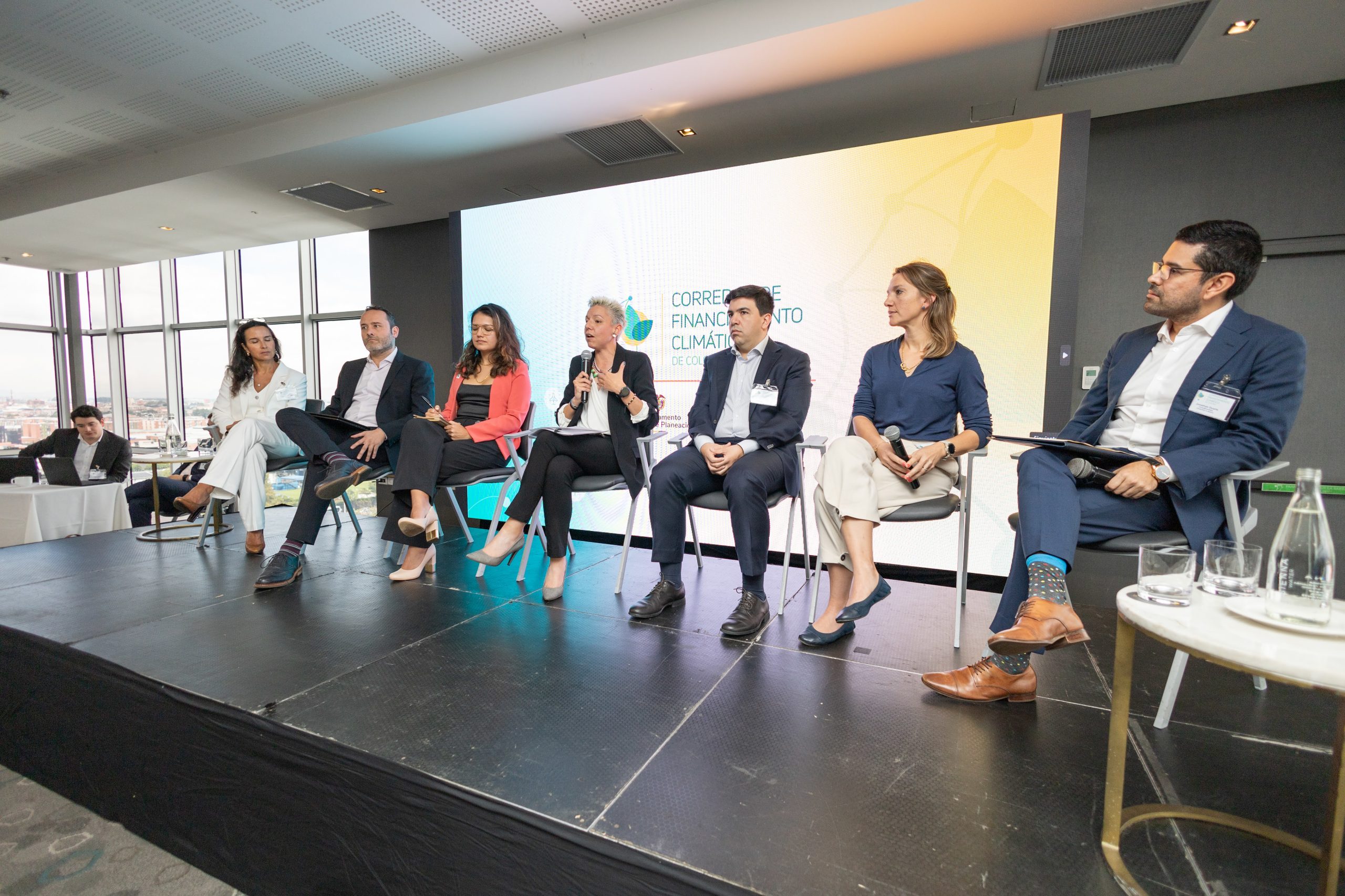 Carolina Díaz Giraldo, DNP Director of Environment and Sustainable Development, speaks during a panel at the "Climate Finance Broker Facility" event in Colombia. She is seated in the center, wearing a white jacket, and engaging in a discussion with the other panelists. The others are listening attentively. In the background, a large screen displays the event's logo.