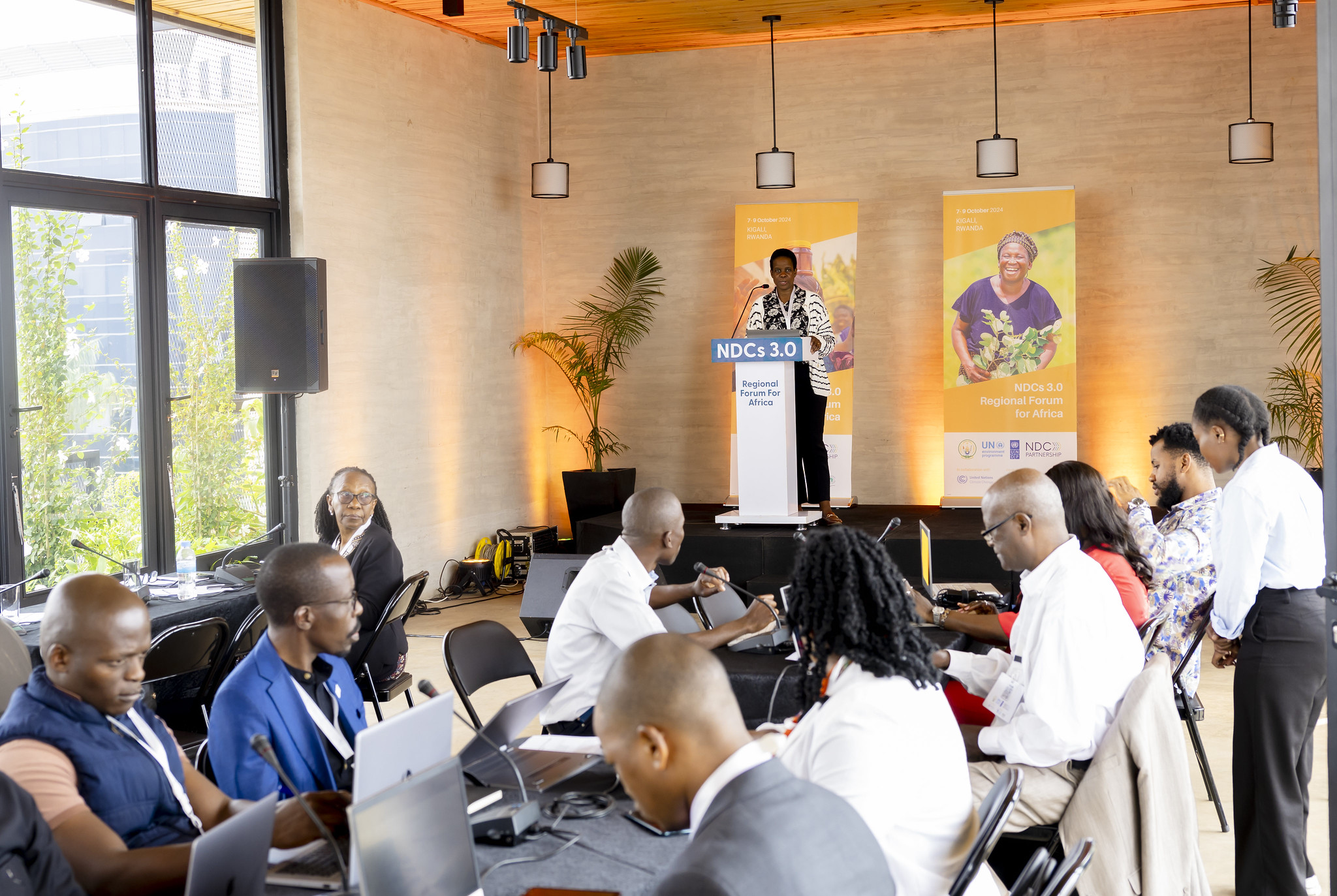 Gloria Namazzi, NDC Partnership Country Engagement Specialist, stands at a podium delivering a speech at the NDCs 3.0 Regional Forum for Africa. Behind her, two banners display the event logo and images of African farmers. In the foreground, a group of people are seated at a table, typing on laptops, engaged in the session.