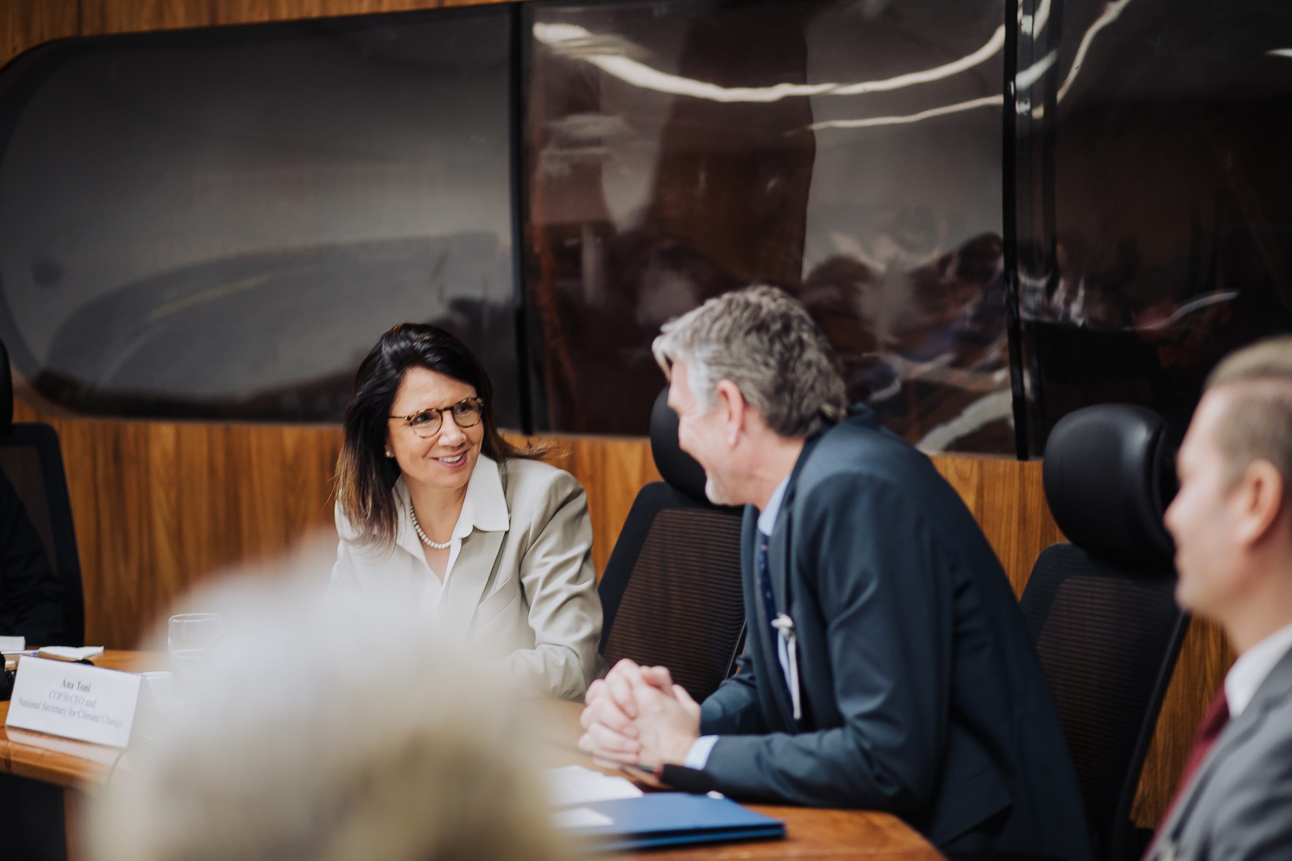 A woman and a man are engaged in a conversation at a meeting, sitting at a conference table. The woman is smiling while listening attentively, and both are dressed in formal attire. The background features a sleek, modern office space with wooden paneling.