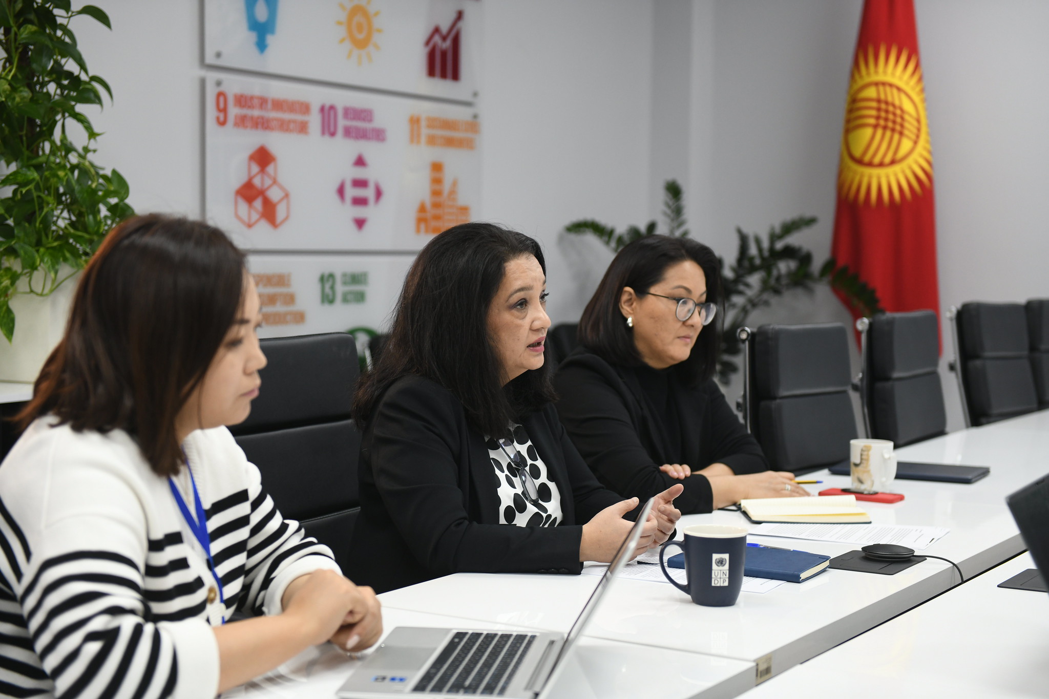 Three women are engaged in a discussion during the NDC Partnership Kyrgyzstan Mission. The woman in the middle is speaking, while the others are listening attentively. They are seated at a conference table with laptops and notebooks in front of them. The background displays icons of the United Nations Sustainable Development Goals, and the Kyrgyzstan flag is visible.