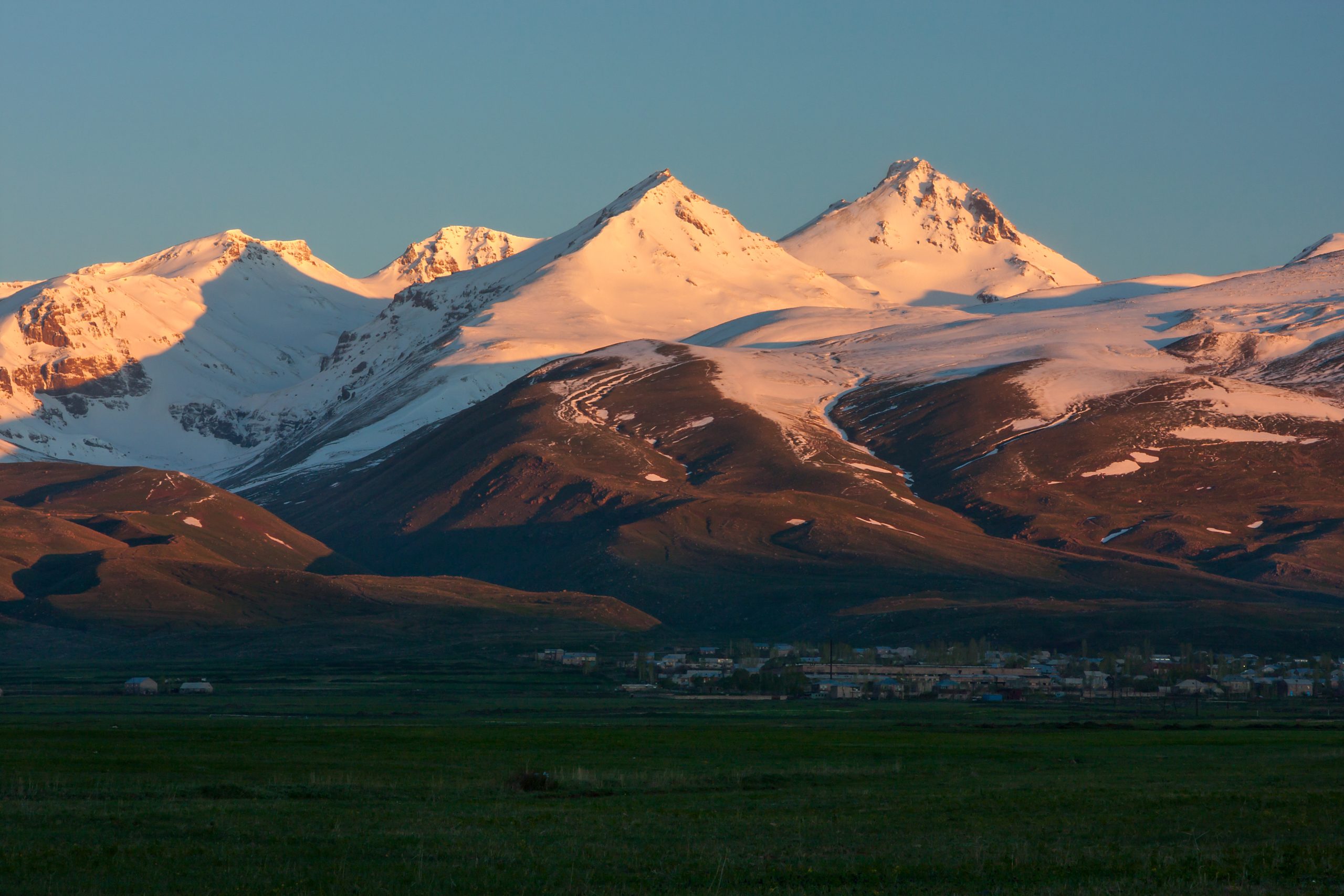 A beautiful view of Aragats, the snow-capped mountain, bathed in the warm rays of sunrise. The sunlight highlights the peaks of the mountain while casting a soft glow over the surrounding slopes. In the foreground, a green landscape transitions into a small town at the foot of the mountain. The peaceful scene captures both natural beauty and human habitation.