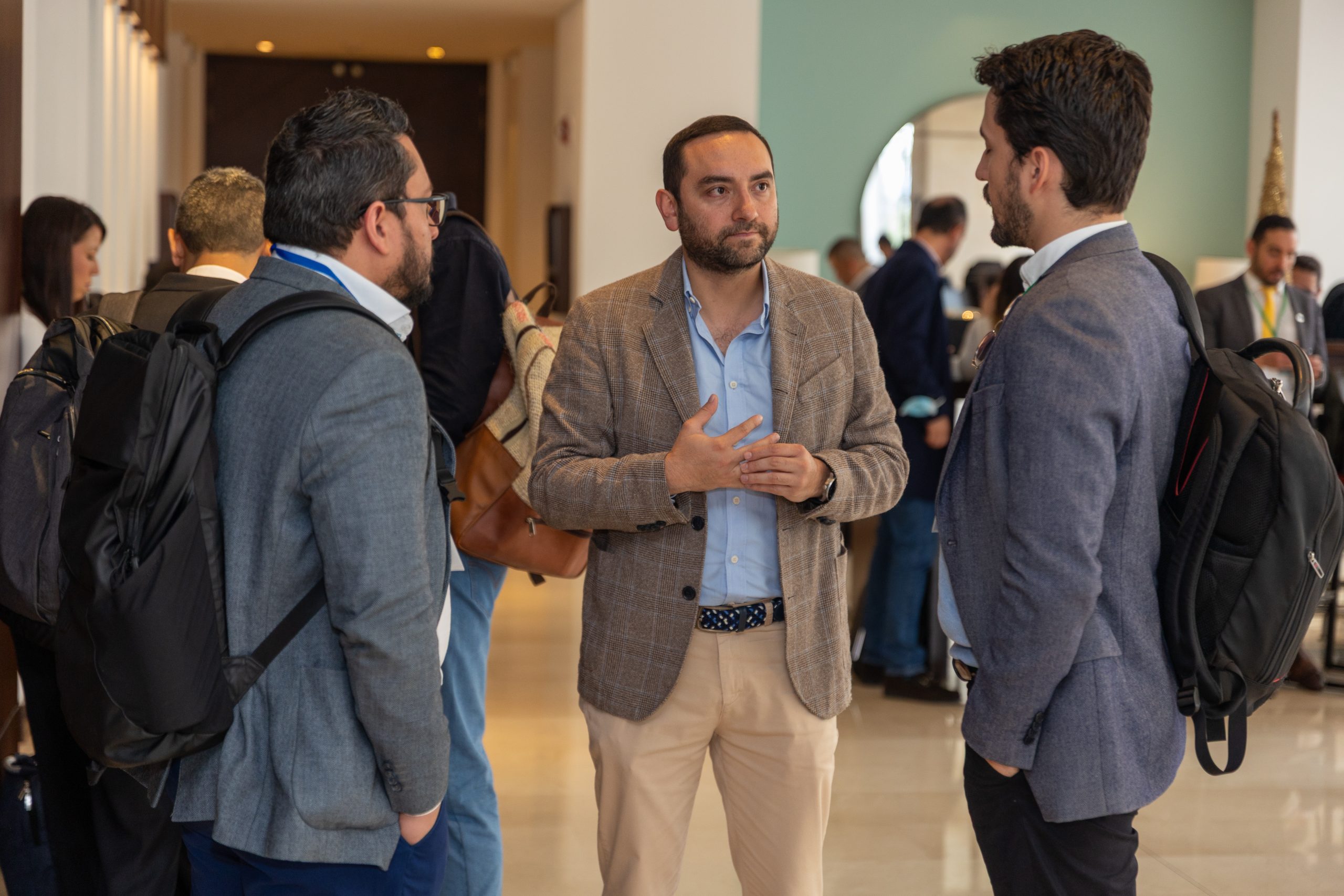 Three professionals are engaged in a conversation during the Climate Finance Broker Facility event in Colombia. One man, wearing a beige jacket and light blue shirt, is speaking while the others listen. They are standing in a spacious, well-lit room with modern décor, and other attendees can be seen in the background.