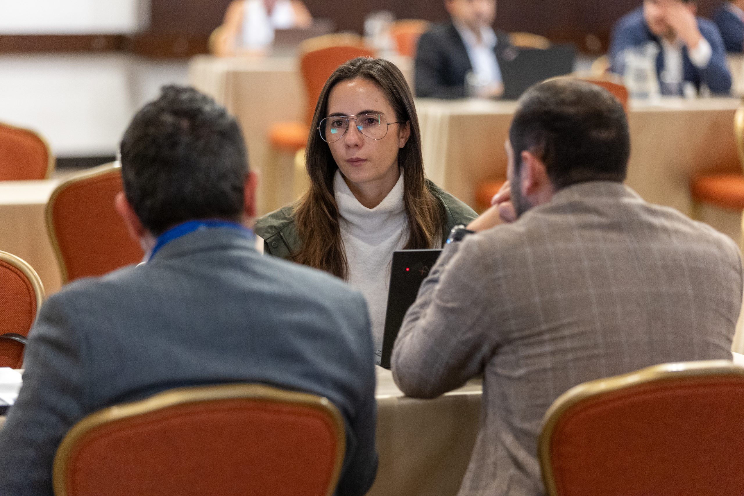 A woman with glasses is engaged in a conversation with two men at the Climate Finance Broker Facility event in Colombia. She is listening attentively, while the men are speaking, with a laptop open in front of them.