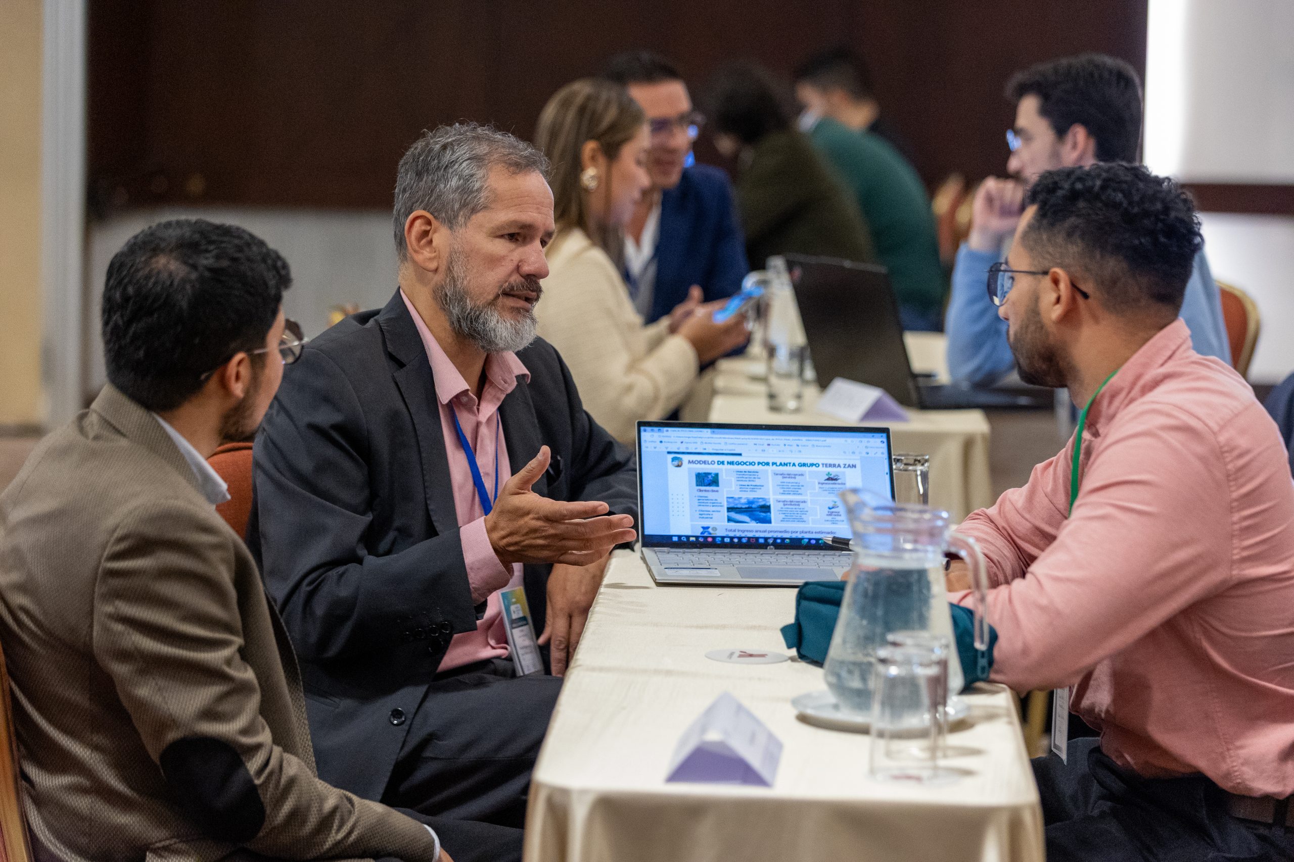 A man with a gray beard is engaged in a conversation with two other professionals at the Climate Finance Broker Facility event in Colombia. One of the men is listening attentively, while the other man is speaking and gesturing. A laptop in front of them displays a document titled "Modelo de Negocio por Planta."