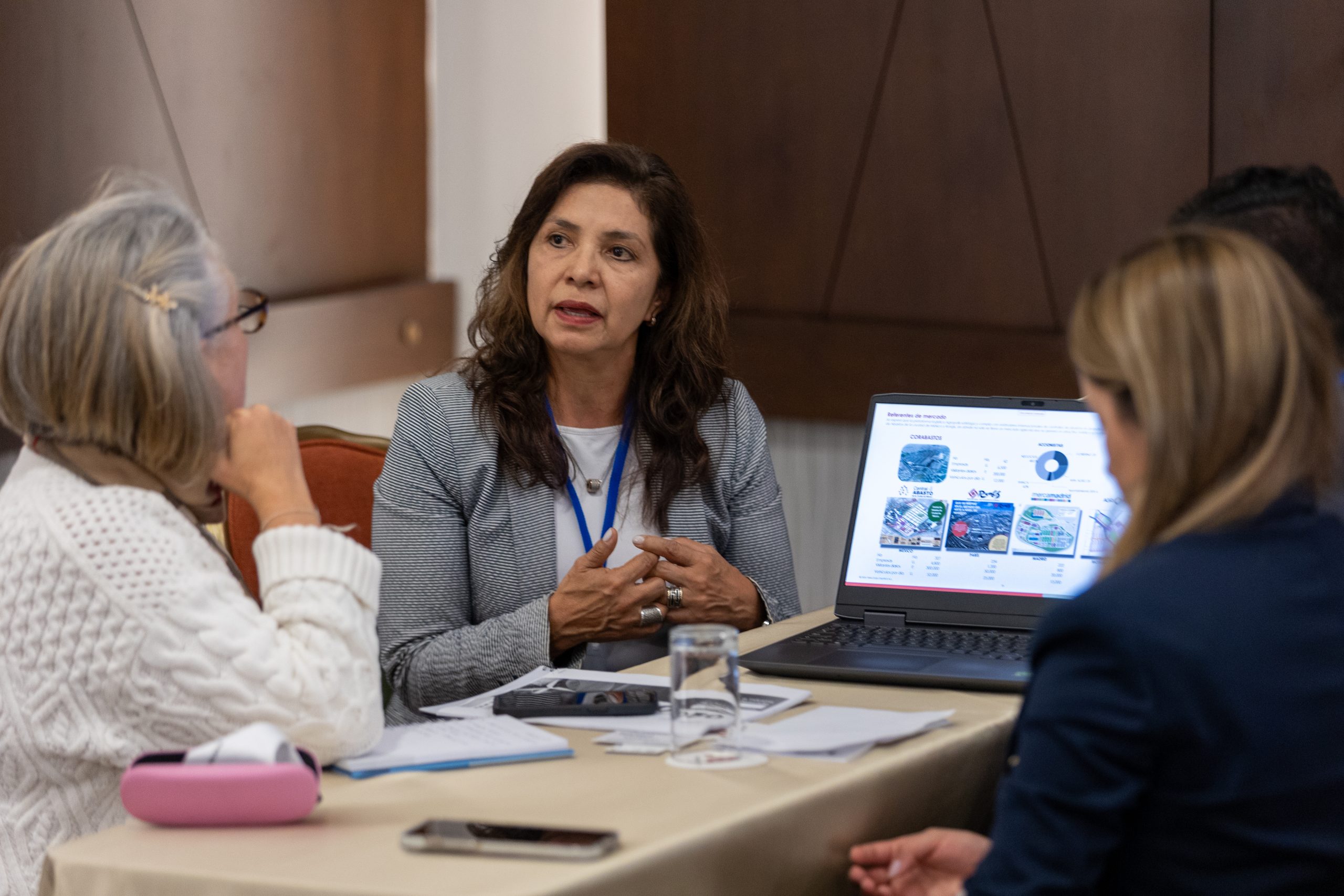 A woman is explaining something during a discussion at the Climate Finance Broker Facility event in Colombia. She is speaking to two other participants seated at the table, with a laptop displaying charts and data beside her. The woman is wearing a gray blazer and a blue ID badge. The participants are engaged in a conversation, with documents and a glass of water on the table.