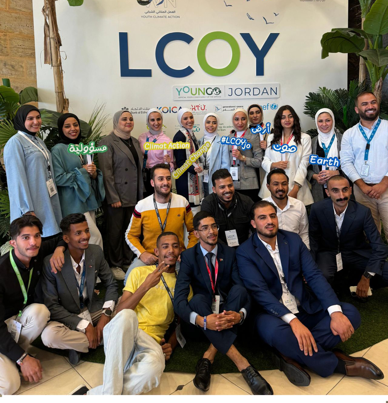 A group of young people pose together for a photo at the LCOY (Local Conference of Youth) event in Jordan. They are standing in front of a backdrop featuring the event logo, which reads "LCOY Jordan," alongside the words "Climate Action" and "Earth." Some individuals are holding colorful speech bubbles with words like "Climate Change" and "#LCOYjo."