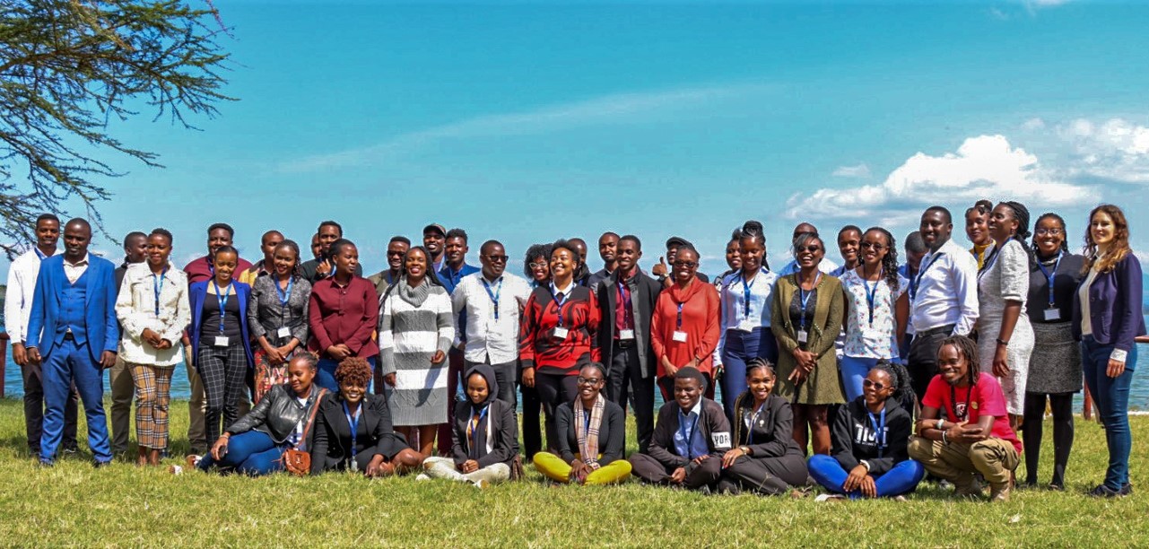 A group photo of young professionals standing and sitting on the grass, smiling and posing for the camera. The group is gathered outdoors, with a bright blue sky and a few clouds in the background. Some individuals are wearing business attire, while others are in casual clothing.